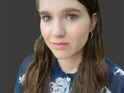A woman with long brown hair wearing a navy blue floral blouse and silver hoop earrings, poses in front of a dark gray background.