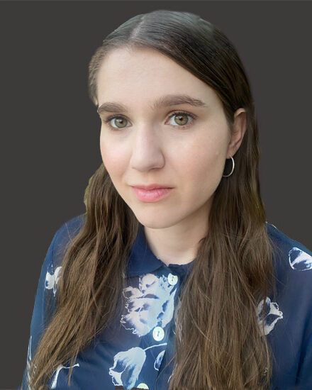 A woman with long brown hair wearing a navy blue floral blouse and silver hoop earrings, poses in front of a dark gray background.